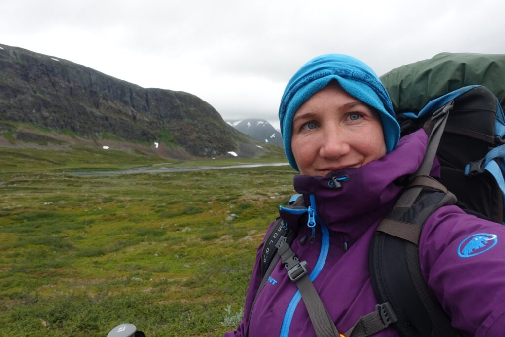 Selfie einer Wanderin in Regenjacke und Mütze, mit schwerem Rucksack in karger, grüner Tundralandschaft.