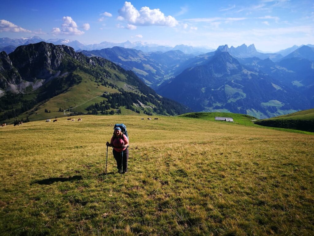 Frau mit Rucksack und Wanderstöcken steigt über eine offene Alpweide, mit weitem Blick auf Berge und Täler.