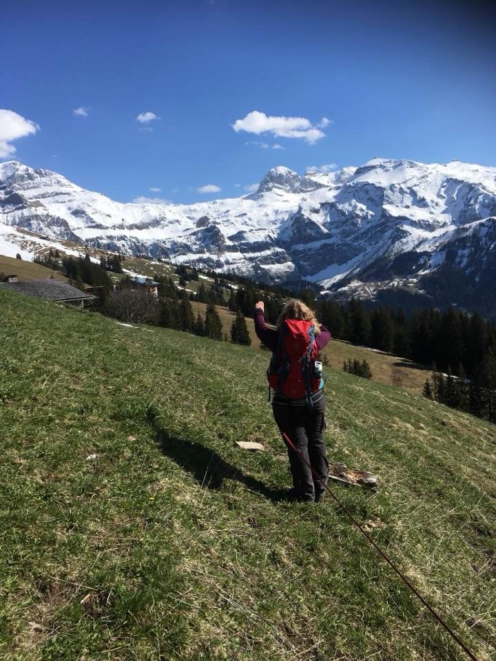 Wanderin mit rotem Rucksack auf einer grünen Wiese, verschneite Bergkette im Hintergrund.