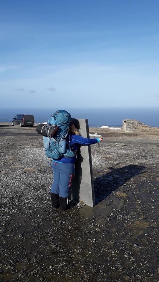 Frau mit großem Trekkingrucksack umarmt einen Grenzstein am Nordkap, im Hintergrund weites Meer und blauer Himmel.