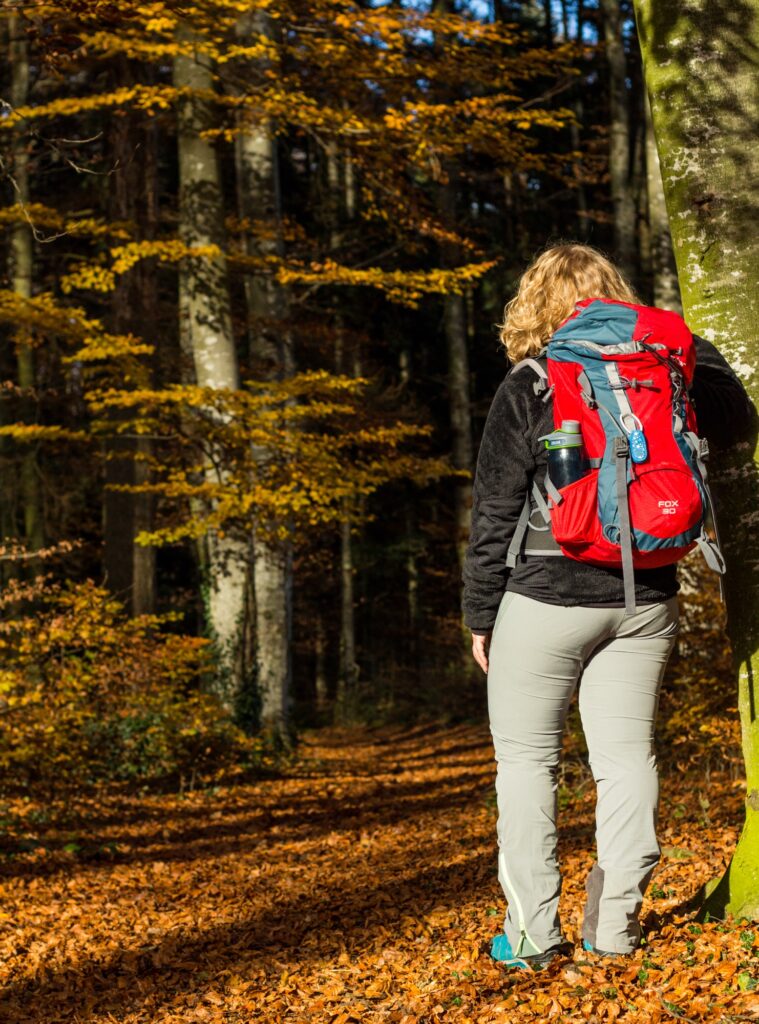 Frau mit rotem Rucksack steht im herbstlichen Buchenwald auf einem Laubteppich.