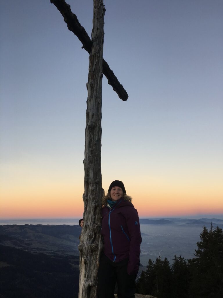 Frau im Winteroutfit lehnt an einem Gipfelkreuz, dahinter Sonnenaufgang über einer weiten Ebene.