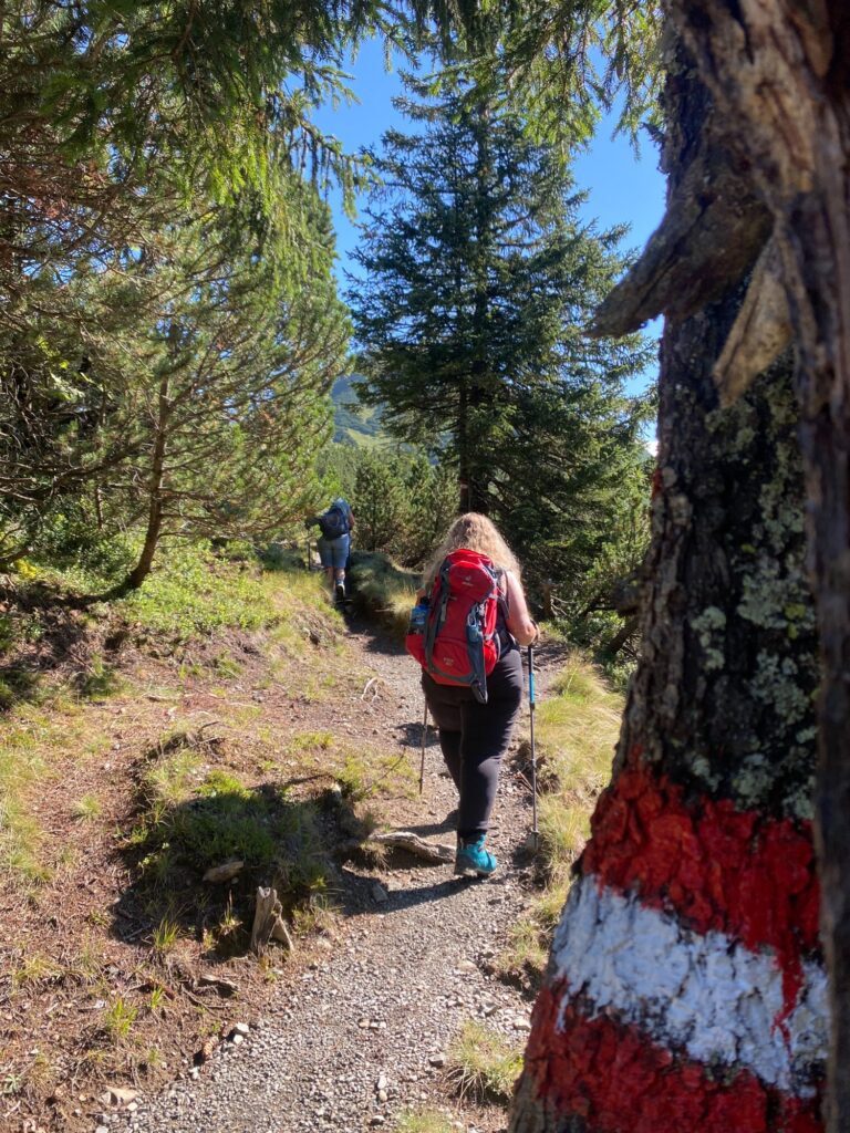 Frau mit rotem Rucksack wandert durch einen Nadelwald, im Vordergrund Baumstamm mit rot-weißer Wegmarkierung.