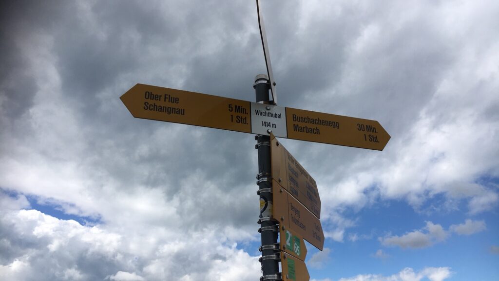 Wegweiser Wachthubel: Gelbe Wanderweg-Schilder auf dem Wachthubel (1414 m) vor grauen Wolken und blauem Himmelsfenster.