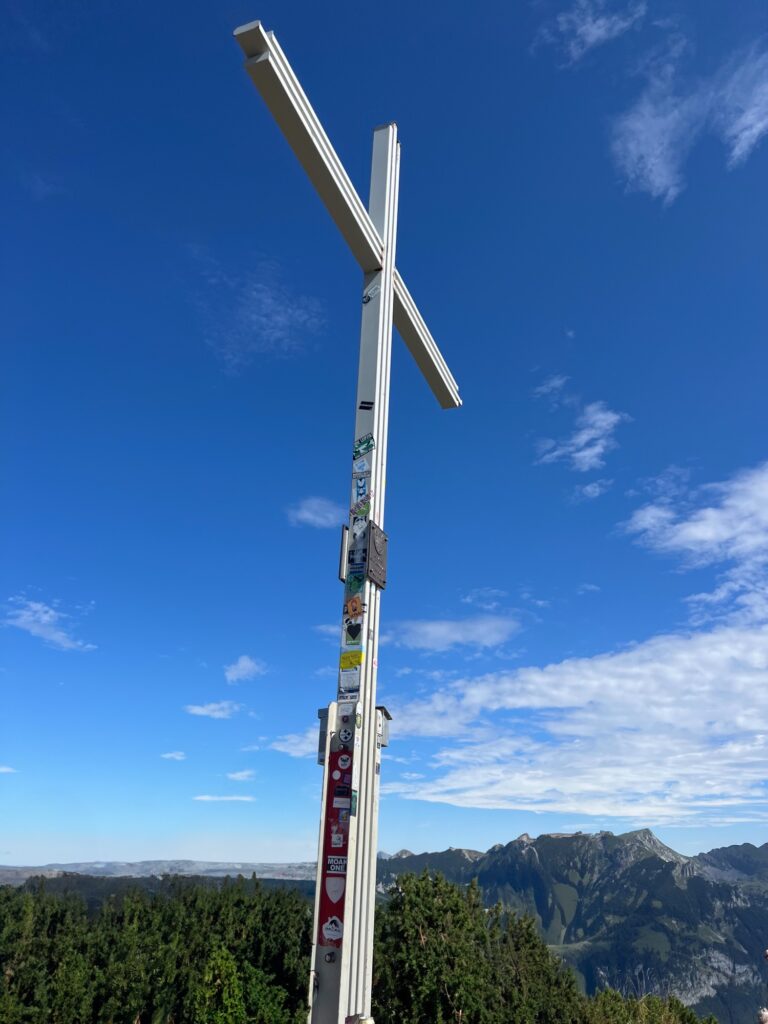 Gipfelkreuz am Bärenkopf: Weißes Metallkreuz mit Stickern, blauer Himmel und Bergpanorama im Hintergrund.