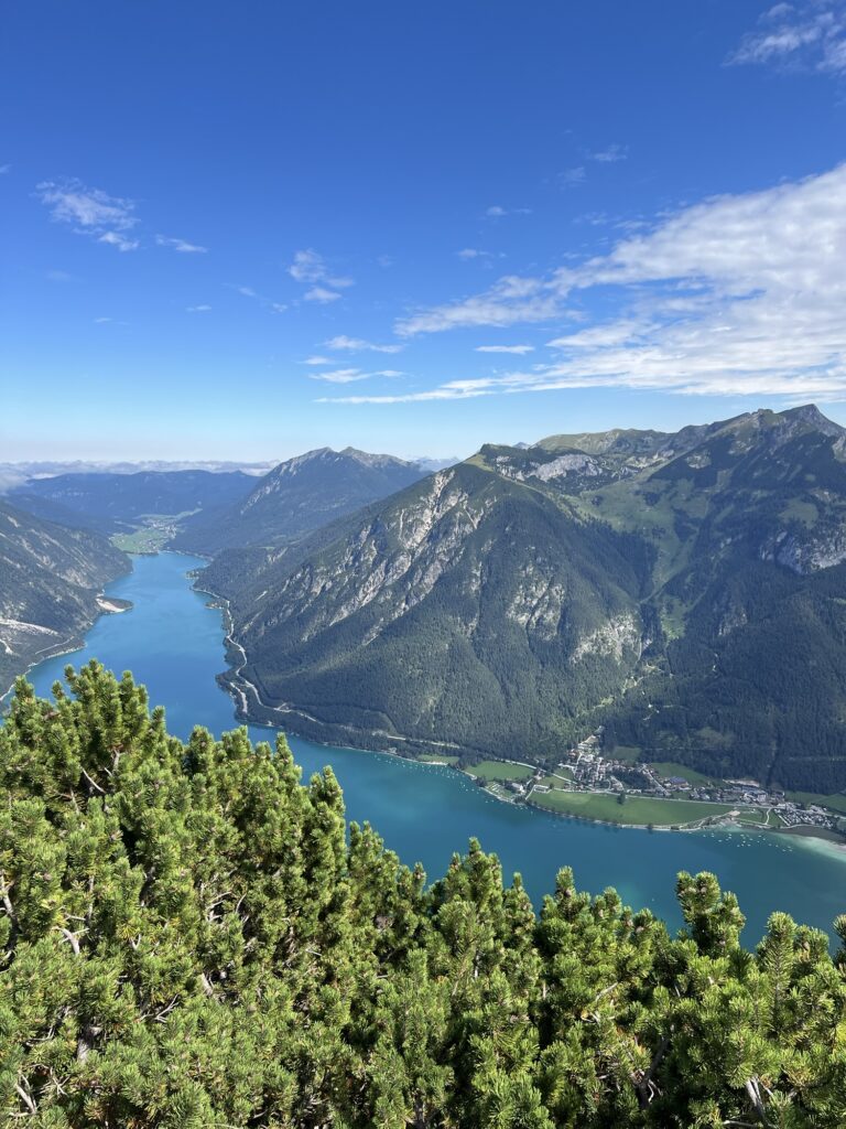 Blick vom Gipfel auf den Achensee: Das türkisblaue Wasser schlängelt sich zwischen den Gebirgsketten, dramatisch unter Wolkenhimmel.