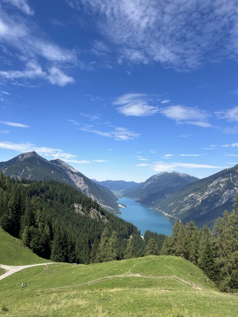 Panoramablick vom Zwölferkopf: Saftig grüne Almwiese im Vordergrund, dahinter der türkisfarbene Achensee eingebettet zwischen hohen Bergen.