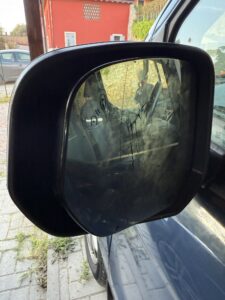Seitenspiegel eines Autos mit feinen Rissen im Glas, im Hintergrund ein rotes Haus und ein parkendes Fahrzeug.