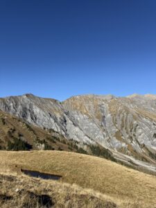 Kleine Wasserfläche inmitten goldener Wiesen – Spiegelung der Felsen und blauer Himmel, ruhige Bergstimmung.