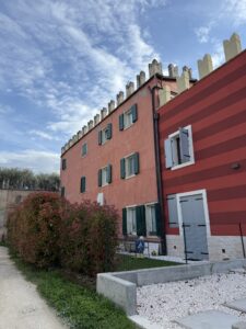 Rotes Wohnhaus mit grünen Fensterläden und Zinnen am Dach, unter blauem Himmel mit Wolken.