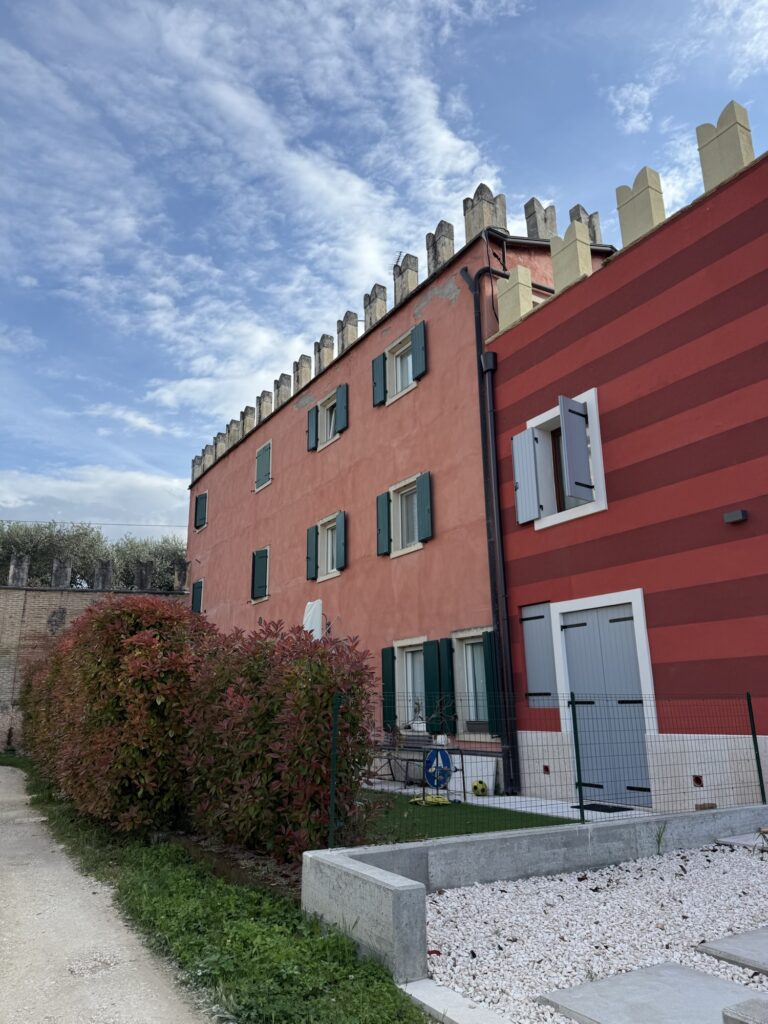 Rotes Wohnhaus mit grünen Fensterläden und Zinnen am Dach, unter blauem Himmel mit Wolken.