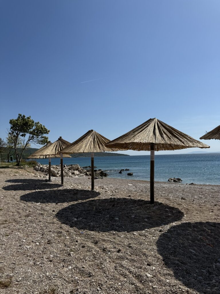 Strand mit Strohdach-Sonnenschirmen und Blick auf das türkisfarbene Meer bei klarem, blauem Himmel.