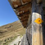 Holzhütte mit gelbem Wanderweg-Schild – traditioneller Unterstand aus Holz am Blumenweg mit Blick auf die Berglandschaft.