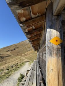 Holzhütte mit gelbem Wanderweg-Schild – traditioneller Unterstand aus Holz am Blumenweg mit Blick auf die Berglandschaft.