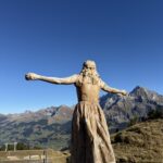 Holzskulptur der Vogel-Lisi mit ausgebreiteten Armen – Symbolfigur am Hahnenmoospass mit Blick auf die Berner Alpen.