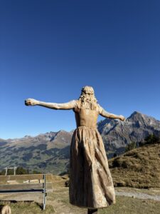Holzskulptur der Vogel-Lisi mit ausgebreiteten Armen – Symbolfigur am Hahnenmoospass mit Blick auf die Berner Alpen.
