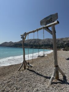 Holzschaukel am Strand von Baška mit türkisfarbenem Wasser und Bergen im Hintergrund, unter strahlend blauem Himmel
