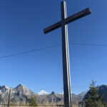 Holzkreuz mit Gipfelblick – schlichtes Kreuz am Blumenweg mit Sicht auf verschneite Alpengipfel und blauen Himmel.
