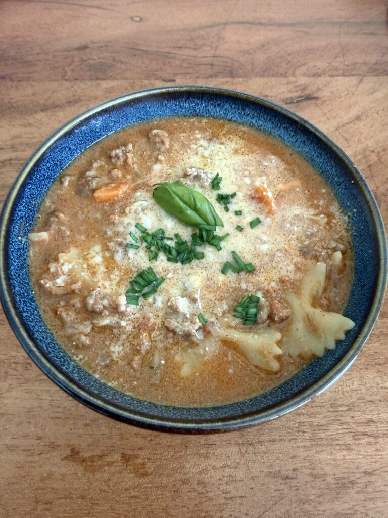 One-Pot Lasagnesuppe mit Farfalle, Rindstatar und Gemüse in einer blauen Schale auf Holztisch – getoppt mit Parmesan, Schnittlauch und Basilikum.