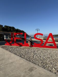 Grosse rote Buchstaben „JELSA“ am Hafen, davor eine Person mit Hund im Sonnenschein.