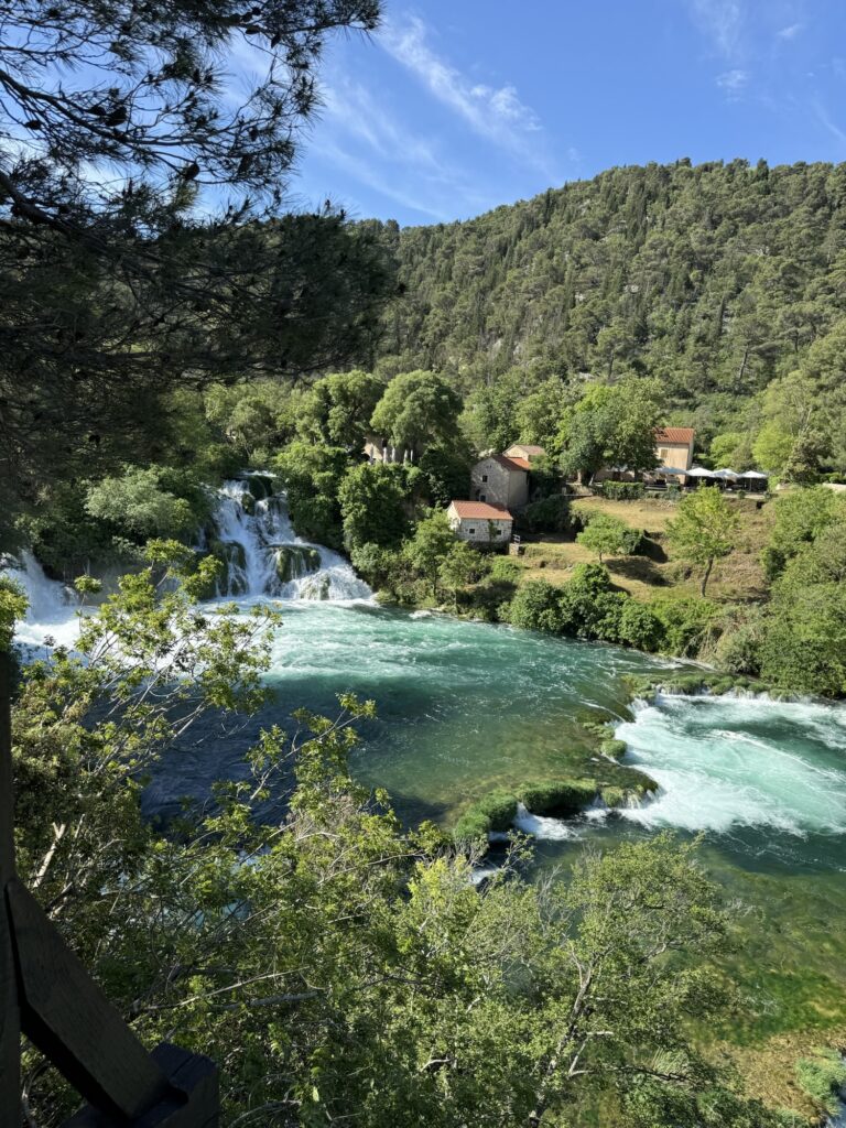 Wasserfall und türkisfarbenes Wasser im Nationalpark Krka, eingerahmt von grünen Bäumen und alten Häusern.