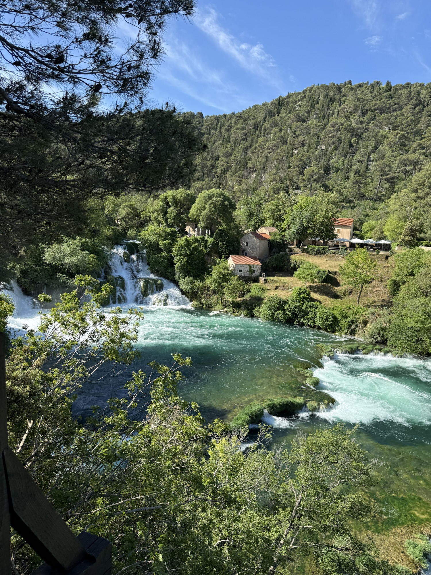 Wasserfall und türkisfarbenes Wasser im Nationalpark Krka, eingerahmt von grünen Bäumen und alten Häusern.