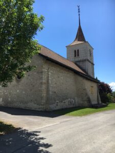 Kirche im Dorf: Alte Dorfkirche aus Stein mit spitzem Turm und Wetterhahn, daneben ein Baum, darüber blauer Himmel.