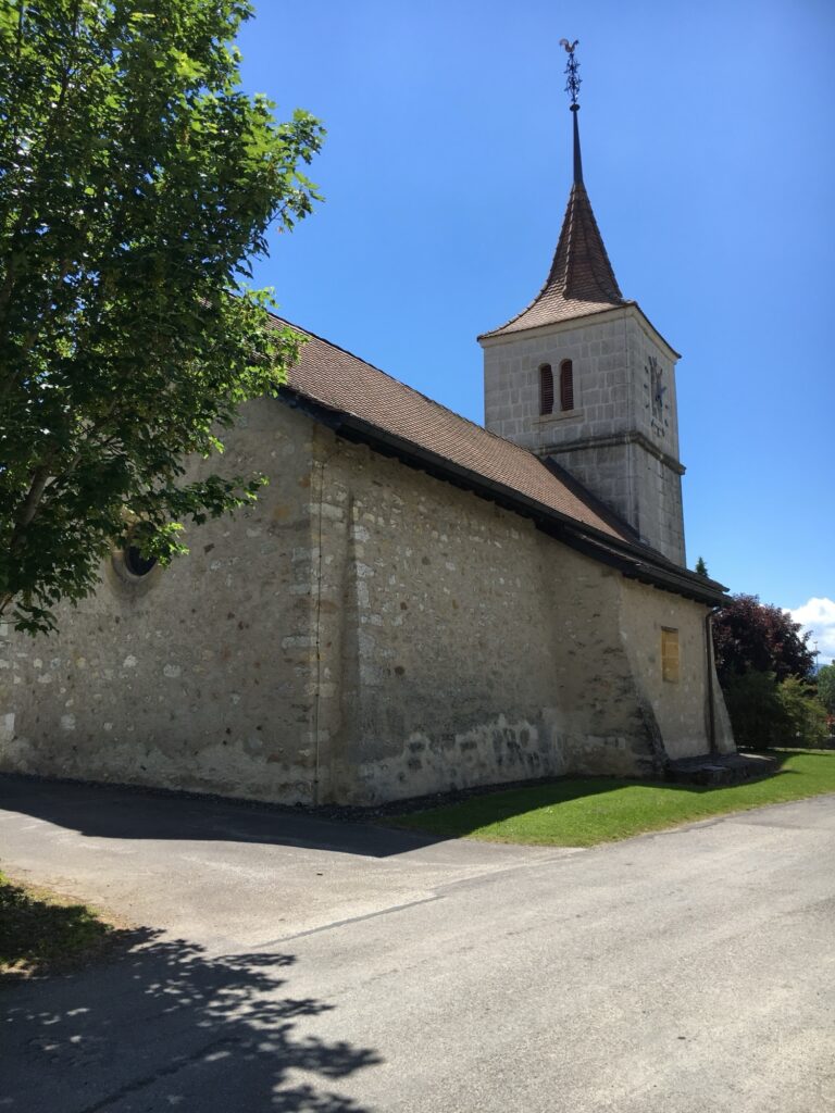 Kirche im Dorf: Alte Dorfkirche aus Stein mit spitzem Turm und Wetterhahn, daneben ein Baum, darüber blauer Himmel.