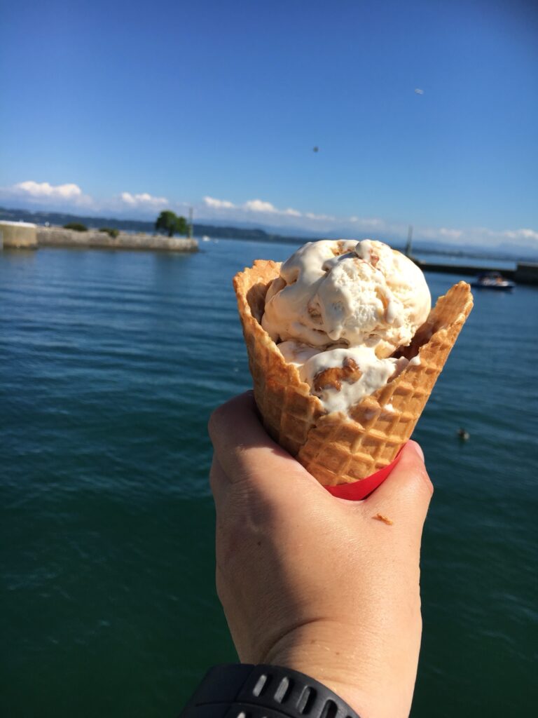Glace am Neuenburgersee: Hand hält eine Waffel mit Eis vor dem blauen Wasser des Neuenburgersees, im Hintergrund Hafenmauer, Boote und Alpen am Horizont.