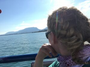 Blick auf den See: Frau mit geflochtenen Haaren und Sonnenbrille sitzt auf einem Boot und schaut über das Wasser zum Jura-Gebirge.