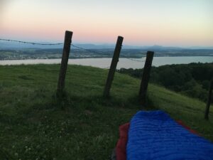 Biwak am See: Schlafsack auf einer Wiese oberhalb des Murtensees, Abenddämmerung mit rosa-blauem Himmel.