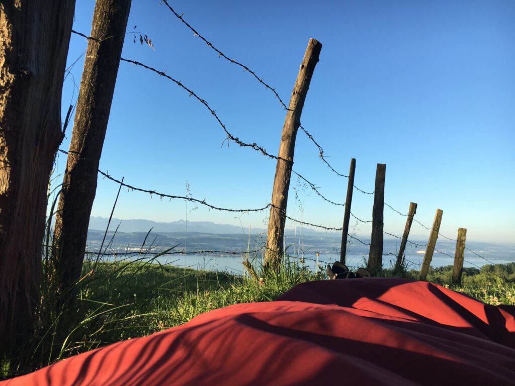 Morgenlicht am Lagerplatz: Schlafsack und Ausrüstung auf einer grünen Wiese mit Blick auf den See und die Alpen im Hintergrund.