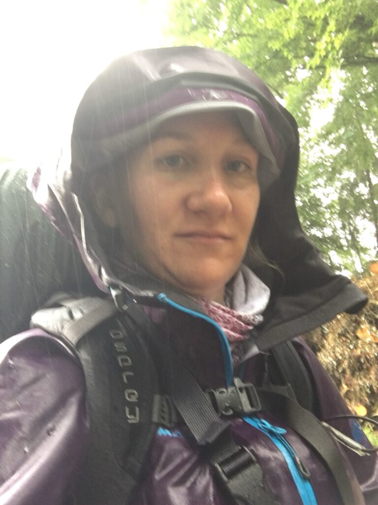 Wanderin im Regen mit Kapuze und Rucksack auf dem Trans Swiss Trail – nass, aber glücklich unterwegs.