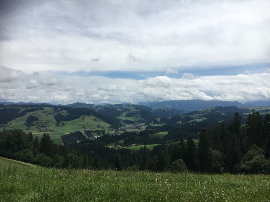 Weite Aussicht über die sanften Hügel des Emmentals mit grünen Wiesen, dunklen Wäldern und Wolkenspiel am Himmel.