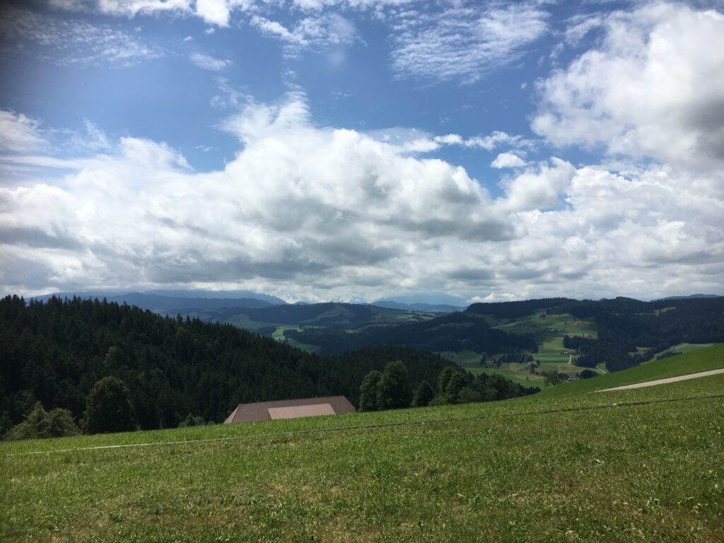 Weite Aussicht über das Emmental – grüne Hügel, Wälder und Bauernhöfe unter einer dramatischen Wolkendecke.