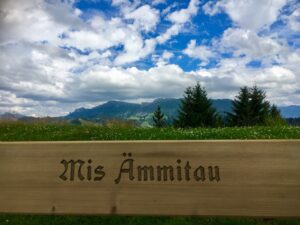 Bank mit Aussicht: Holzbank mit der Aufschrift „Mis Ämmitau“, dahinter weite Sicht auf Hügel und Berge unter dramatischem Himmel.