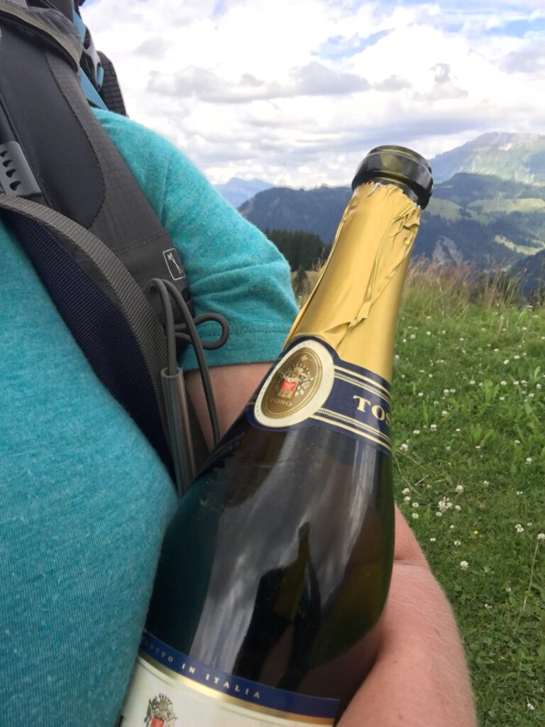 Flasche unterwegs: Wanderin mit Rucksack trägt eine leere Champagnerflasche am Berg, aufgehoben zur späteren Entsorgung.