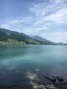 Sarnersee Ufer: Blick vom Kiesstrand auf das türkisfarbene Wasser des Sees, dahinter grüne Hügel.