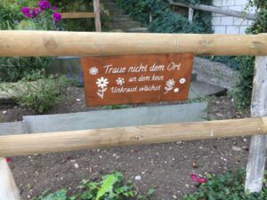 Gartenspruch: Schild mit der Aufschrift „Traue nicht dem Ort an dem kein Unkraut wächst“ an einem Holzzaun mit Blumen im Hintergrund.