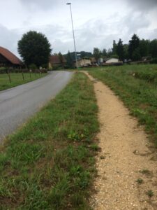 Schmaler Kiesweg neben einer Landstrasse mit Bauernhäusern im Hintergrund bei grauem Regenwetter.