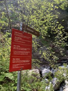 Wegweiser zum Jungbrunnen – Rotes Hinweisschild mit Sicherheitshinweisen vor einem Bachlauf im Grünen.