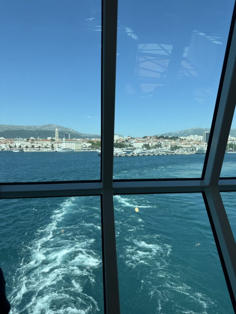 Blick durch grosse Fenster einer Fähre auf die Hafenstadt Split in Kroatien mit ihrer Uferpromenade, historischen Gebäuden und dem tiefblauen Meer.