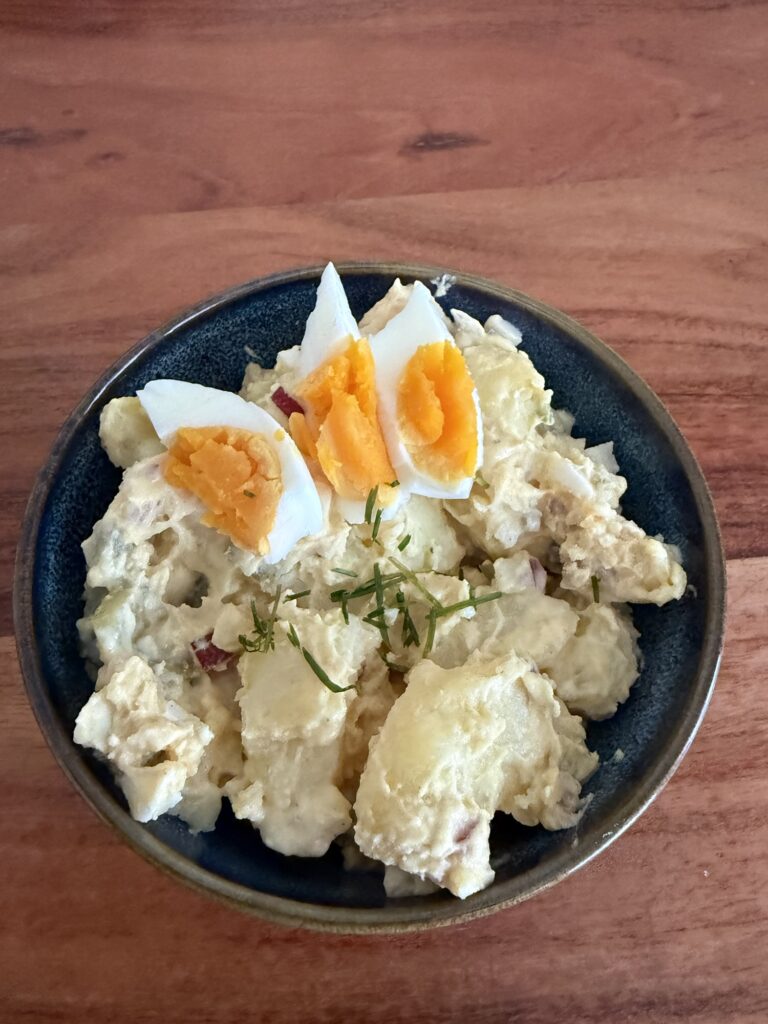 Cremiger High-Protein-Kartoffelsalat mit Eiern als Topping, serviert in einer Schale auf einem natürlichen Holzhintergrund.