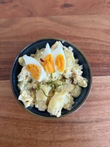 High-Protein-Kartoffelsalat in einer dunkelblauen Schale, garniert mit hartgekochten Eiern und frischen Kräutern auf einem Holztisch.
