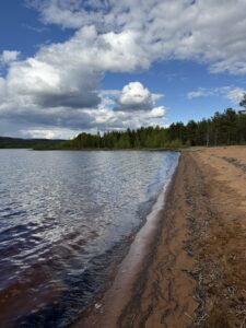 Sandstrand am See in Schweden – Weiches Licht über dem See, ruhige Wellen treffen auf den rötlichen Sandstrand, umgeben von Wäldern und weitem Himmel.