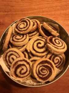 Runde Dose gefüllt mit selbstgemachten Zimtschnecken-Plätzchen: goldbraune Kekse mit deutlich sichtbarer Zimt-Zucker-Spirale, dicht aneinandergeschichtet auf Backpapier, aufgenommen von oben auf warmem Holztisch.