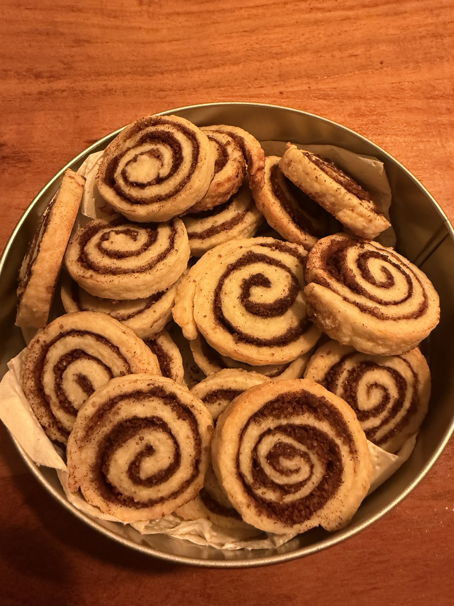 Runde Dose gefüllt mit selbstgemachten Zimtschnecken-Plätzchen: goldbraune Kekse mit deutlich sichtbarer Zimt-Zucker-Spirale, dicht aneinandergeschichtet auf Backpapier, aufgenommen von oben auf warmem Holztisch.