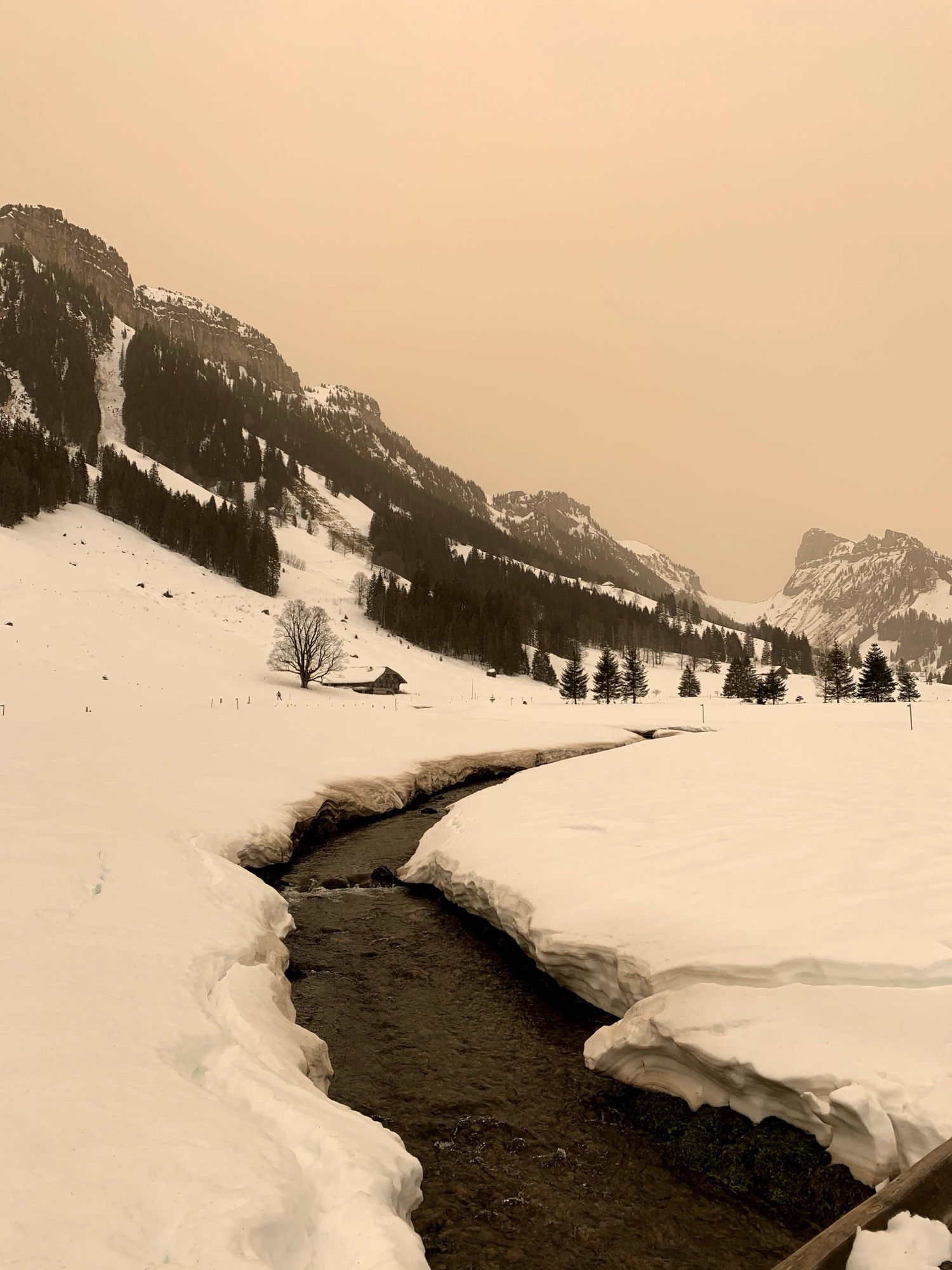 Tiefer Bachgraben im Schnee im Justistal, dahinter verschneites Tal mit Alphütten und steilen Bergflanken.