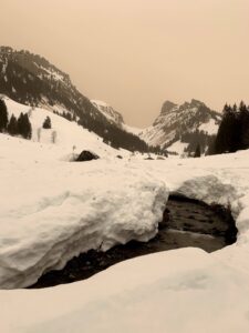 Winterlicher Bach im Justistal, der sich zwischen hohen Schneebänken durchzieht, umgeben von Wald und Felswänden.
