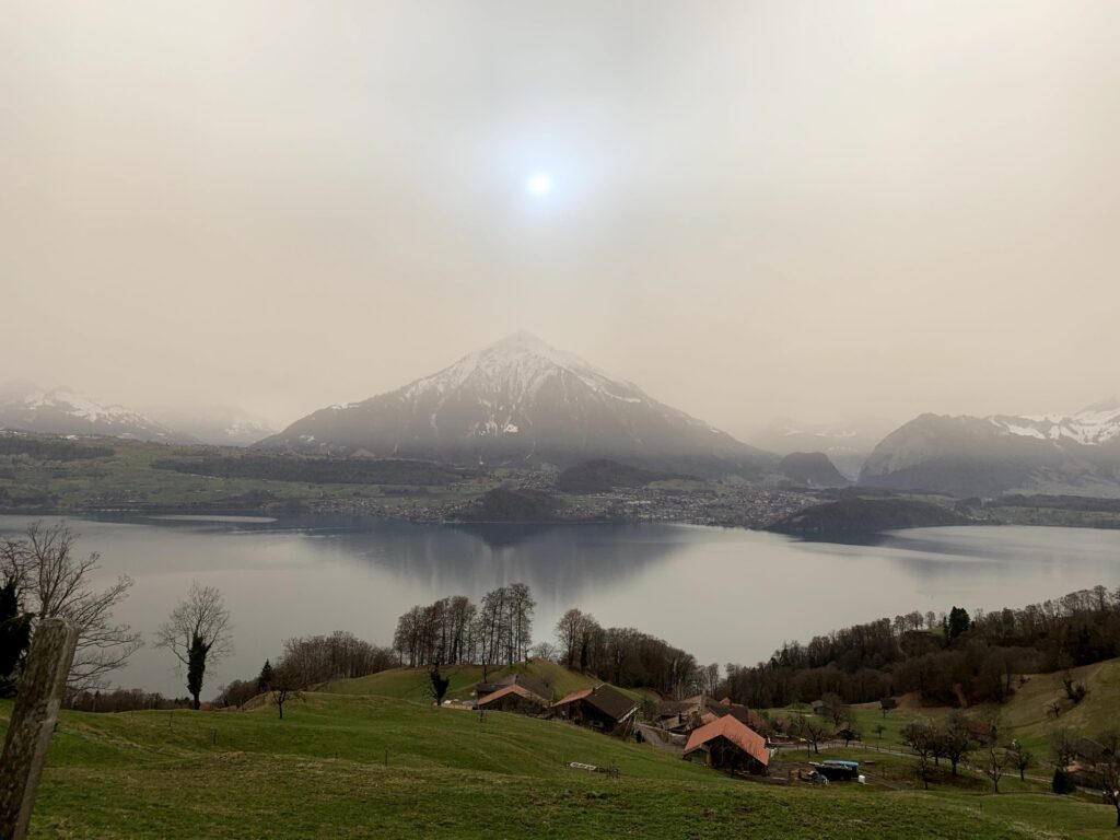 Blick vom Justistal auf den Thunersee mit sanften Hügeln, Bauernhöfen und Nebel über dem Wasser im Berner Oberland.